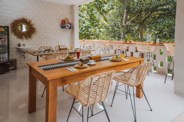 Outdoor tropical breakfast table with fresh fruits, salads, eggs, and juices on a wooden surface, rattan chairs, white brick balcony, and lush green backdrop. - Bali Villas